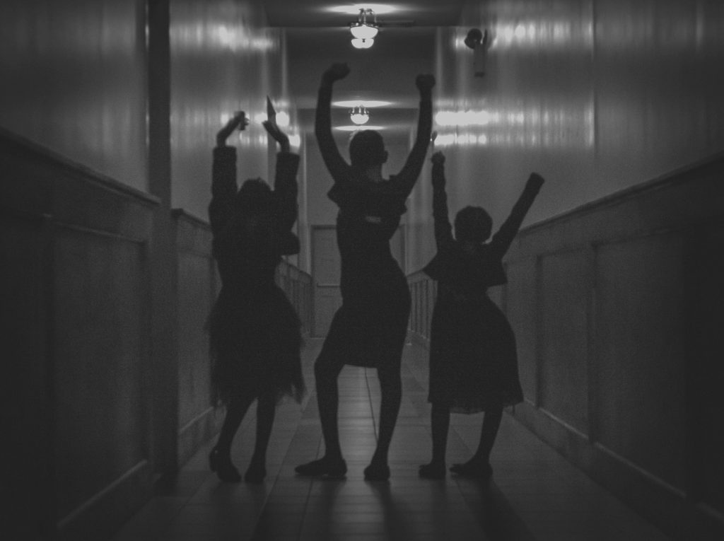 Black-and-white image of three young girls in a church hallway raising their hands in the air.