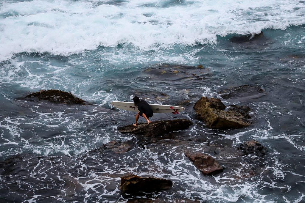 A woman with a surfboard climbs on the rocks by the ocean.